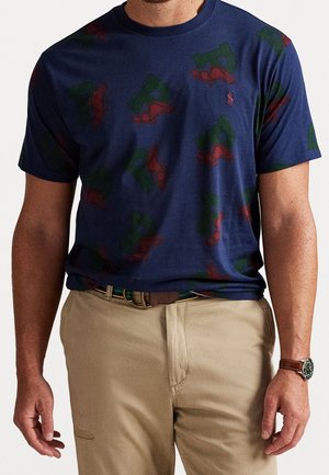 Mand iført navyblå T-shirt med grønne og røde skologoer, beige bukser, brunt bælte og armbåndsur, stående mod hvid baggrund.