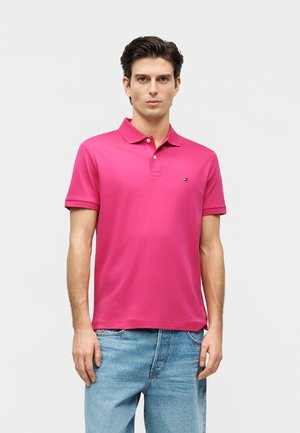 Uomo con polo rosa acceso a maniche corte e jeans in denim azzurro chiaro, in piedi su uno sfondo bianco uniforme.