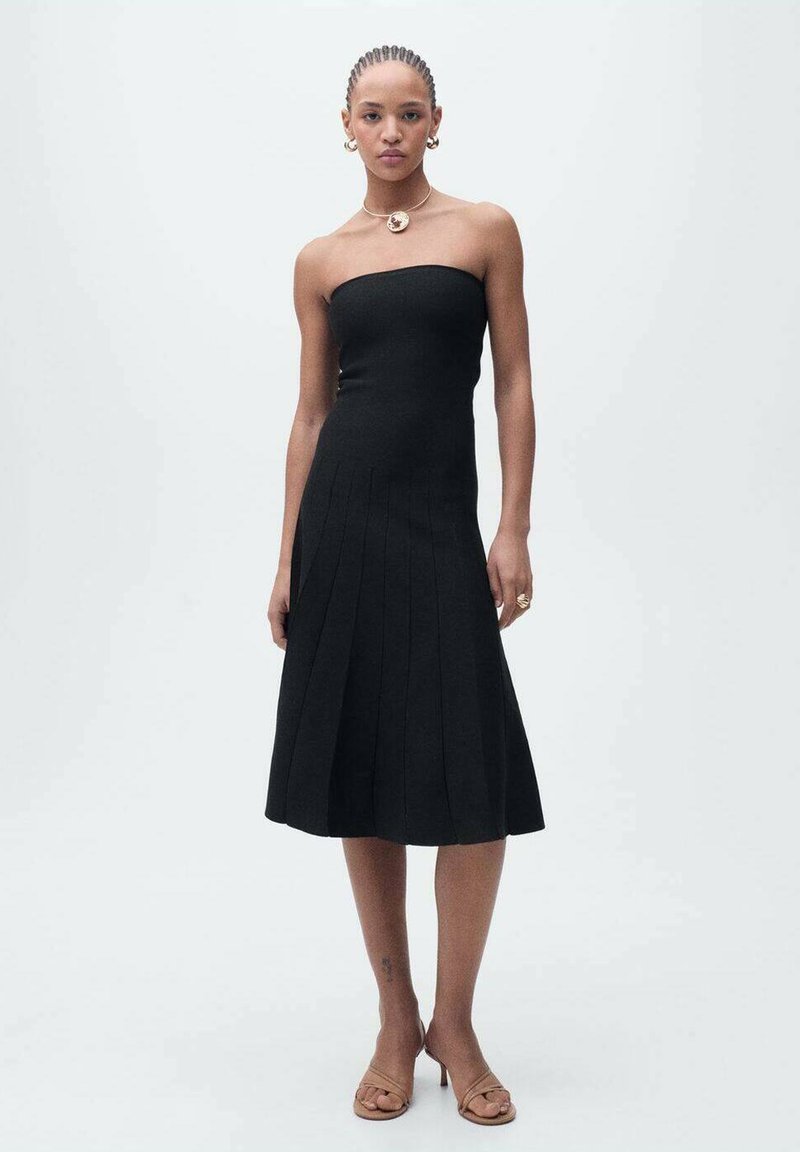 Rochie neagră fără bretele, dintr-un material ribat, având o fustă evazată cu plii verticale. Accesorizată cu bijuterii minime și sandale neutre.