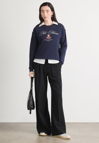 Granatowy sweatshirt z białym haftowanym napisem, nałożony na biały kołnierzyk. Czarne spodnie o szerokich nogawkach i buty z srebrnymi akcentami dopełniają stylizację.
