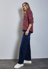 Haut bordeaux à manches longues avec des motifs abstraits blancs, associé à un jean large bleu foncé et des baskets blanches, sur un fond neutre.