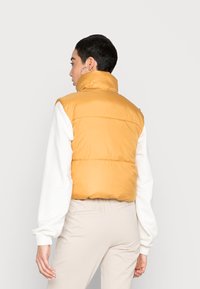 Colete acolchoado amarelo com gola alta, design quilted e acabamento brilhante, usado sobre uma camisa de manga longa branca.
