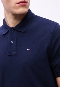 Κοντινό πλάνο ενός ατόμου που φοράει μπλε σκούρο πόλο μπλουζάκι με δύο κουμπιά και ένα μικρό κόκκινο, λευκό και μπλε λογότυπο στο στήθος.