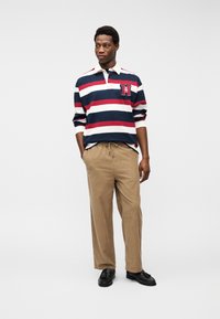 Νέος άνδρας που φοράει μπλε σκούρα, κόκκινη και άσπρη ραβδωτή ράγκμπι μπλούζα με σήμα, μπεζ παντελόνια και μαύρα λοφωτά παπούτσια με φούντες, στέκεται με τα χέρια στις τσέπες.