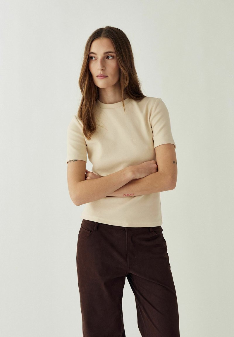Femme aux longs cheveux bruns portant une chemise beige à manches courtes et un pantalon marron foncé, debout les bras croisés, regardant vers la gauche.