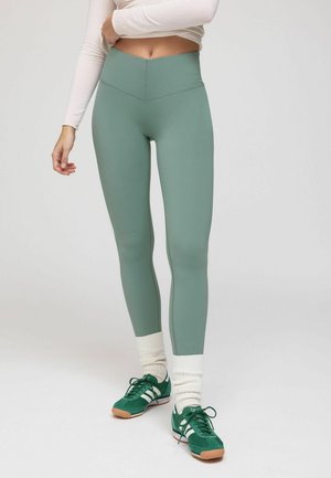 Nő zsályazöld magas derekú leggingsben, fehér hosszú ujjú felsőben, krémszínű lábszármelegítőben és zöld Adidas tornacipőben áll egyszínű háttér előtt.