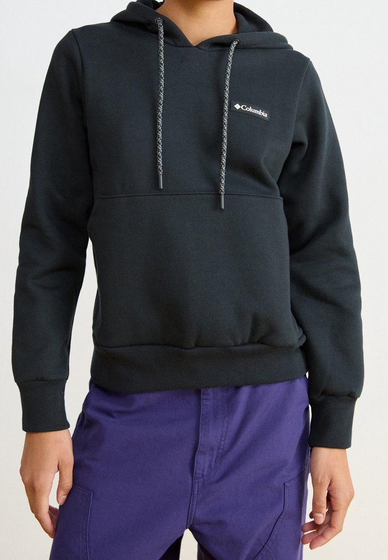 Persoon gekleed in een zwarte Columbia-hoodie met patroon op de trekkoorden en paarse broek, staand met handen ontspannen langs het lichaam.