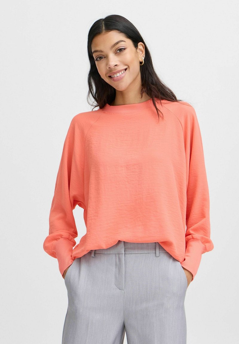 Top a maniche lunghe color corallo con vestibilità rilassata, scollatura a coste e una texture delicata, abbinato a pantaloni grigi chiari con un taglio sartoriale.