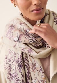 Bufanda de flores en color beige con intrincados estampados botánicos en púrpura, confeccionada en un tejido suave, drapeada elegantemente alrededor del cuello. Anillo dorado visible en la mano.