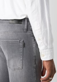 Jeans de mezclilla gris con una textura suave, diseño clásico de cinco bolsillos y una etiqueta de marca de cuero en la parte trasera de la cintura. Camisa blanca visible.