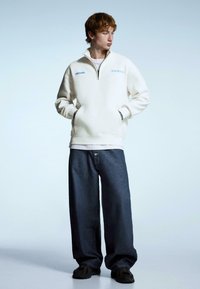 White fleece half-zip pullover with front pockets, paired with loose dark denim jeans and black sneakers. Simple, functional design.