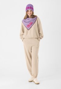 Beiges Loungewear-Set mit elastischen Bündchen, lila gestrickter Schal mit Streifenmuster und roter Schrift, passender gestreifter Beanie, lässige Sneaker.