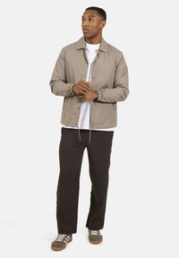 Jasnobrązowa kurtka z kołnierzem, zatrzaskami i elastycznymi mankietami, noszona na białej koszuli i czarnych spodniach, zestawiona z szarymi tenisówkami.