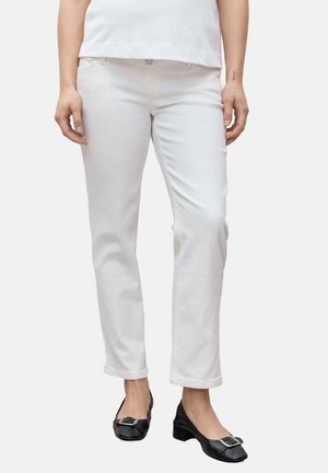 Vrouw draagt witte rechte broek en zwarte lage hakken met vierkante zilveren gespen, staand tegen een effen achtergrond.