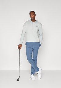 Moleton de manga longa cinza claro com um bolso frontal, combinado com calças azuis e ténis brancos. Um taco de golfe é segurado na mão direita.