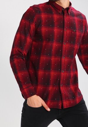 Punainen ja musta ruudullinen paita, jossa on napillinen kaulus, pieniä valkoisia pilkkuja ja pitkät hihat; valmistettu pehmeästä, kudotusta kankaasta.