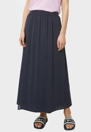 Femme portant une jupe noire longueur cheville avec une taille élastique, un haut rose clair et des sandales noires ouvertes à l'avant avec des inscriptions blanches.
