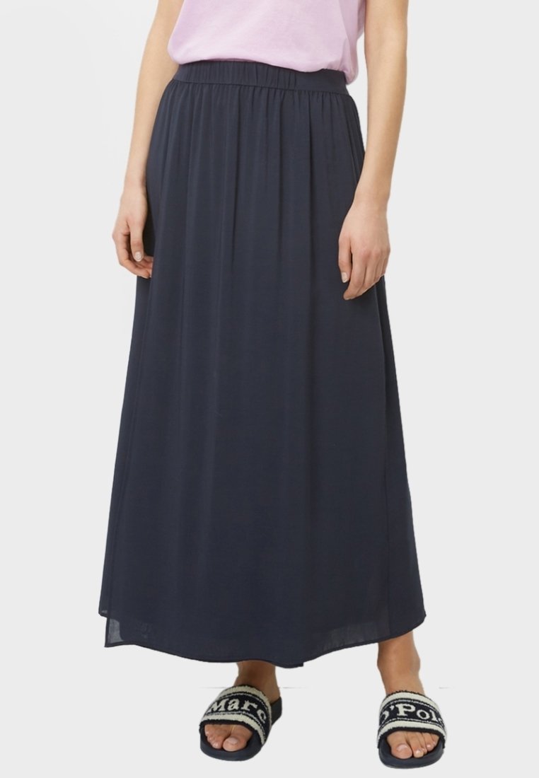 Femme portant une jupe noire longueur cheville avec une taille élastique, un haut rose clair et des sandales noires ouvertes à l'avant avec des inscriptions blanches.