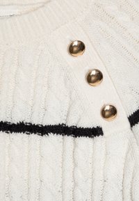 Suéter blanco de punto trenzado con una franja horizontal negra y tres detalles de botones dorados en el hombro.