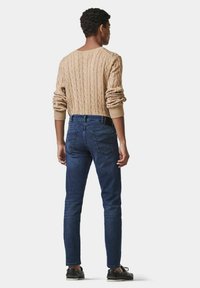 Pull en maille beige avec des poignets côtelés, associé à un jean foncé ajusté et des chaussures noires, vu de dos sur fond blanc.