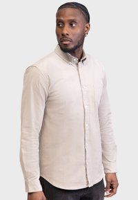 Lys beige skjorte med knapper og lange ærmer, brystlomme og et tekstureret stof, som har en rund kant og en krave.
