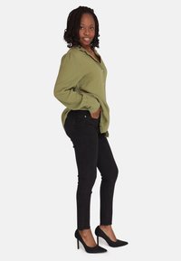 Camisa de botones de manga larga en un verde oliva claro; jeans ajustados negros y zapatos de tacón alto negros; cabello ondulado y pose relajada.