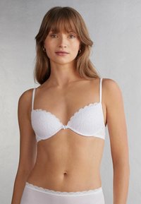 Bílá krajková podprsenka se vlnitými okraji, hlubokým výstřihem a nastavitelnými tenkými ramínky. Na košíčkách jsou jemné květinové vzory.