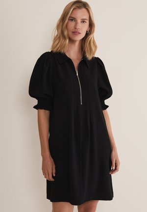 Vrouw met schouderlang blond haar, gekleed in een zwarte jurk tot op de knie met pofmouwen en een rits aan de voorkant, staand tegen een effen achtergrond.