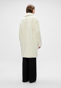 Palton din blană artificială de culoare crem, cu o croială lejeră, guler lat și mâneci lungi. Purta pe deasupra unor pantaloni negri cu croială largă, care au o suprafață texturată.
