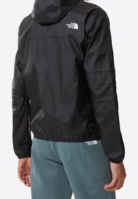 Person som bär en svart huva vindjacka och turkosa fleecbyxor, båda med vita The North Face-logotyper, visad bakifrån.