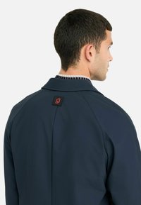 Giacca blu navy con colletto strutturato, caratterizzata da una rifinitura testurizzata e un logo del marchio ben visibile sul retro. Tessuto liscio, design minimale.