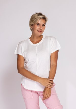 Woman with short blonde hair wearing a white short-sleeve shirt and light pink pants, standing against a plain light background.