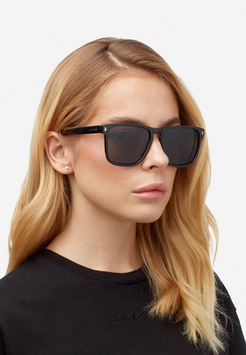Črne pravokotne sončne očala s temnimi lečami, z mat površino in minimalističnim oblikovanjem. Nosijo jih oseba z dolgimi blond lasmi.