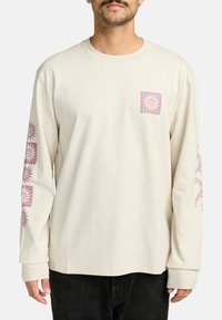 Camicia a maniche lunghe di colore crema in cotone con una stampa di un sole sulla tasca anteriore. Presenta grafiche simili del sole lungo le maniche.