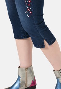 Jeansowe rybaczki w ciemnym niebieskim kolorze z haftowanymi kwiatami z boku; zestawione ze metalicznymi botkami za kostkę w srebrnym, różowym i niebieskim kolorze z zamkami błyskawicznymi.