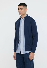 Cardigan zippé bleu marine avec un col haut, fabriqué en tissu doux. Détail avec un logo sur la poitrine et associé à une chemise rayée en dessous.
