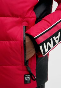 Rote gepolsterte Jacke mit einem schwarzen Ärmel und weißer Schrift, Reißverschlusstasche auf der linken Seite und einem Etikett mit "Camp David"-Markierung.