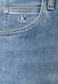 Světle modrá džínová tkanina s přední kapsou a bílým vyšívaným logem "CK" a zlatými švy. Texturovaný, pohodlný design.