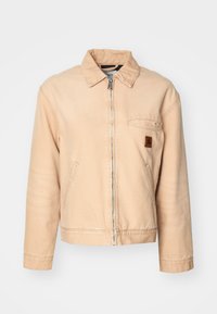 Giacca in denim beige con zip frontale, collo classico, tasca sul petto e due tasche laterali. Presenta cuciture sottili e una toppa con logo in pelle.