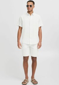 Camisa blanca de manga corta con botones, combinada con pantalones cortos a juego, ambos hechos de tela ligera. Sandalias marrones y gafas de sol completan el conjunto.