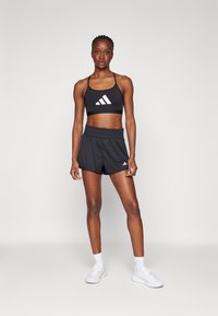 Svart atletisk crop-top med hvit logo, kombinert med svarte shorts med samlet midjebånd. Modell iført hvite sneakers og sokker.