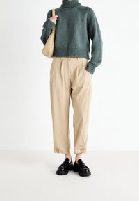 Personne portant un pull à col roulé vert olive, un pantalon beige taille haute raccourci avec des boutons, des mocassins noirs, et tenant un sac à main beige.