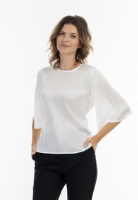 Fehér, könnyű blúz 3/4-es harangujjakkal, sima textúrával, kerek nyakkivágással és finom függőleges csíkokkal. Fekete nadrággal viselve.