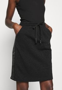 Falda negra con un acabado texturizado y brillante. Presenta una cintura con cordón y bolsillos laterales. Tiras de material negro liso en los bordes.