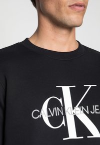 Černý bavlněný svetr s kulatým výstřihem, žebrovanými lemy a velkým bílým logem "CK" s textem "CALVIN KLEIN JEANS" pod ním.