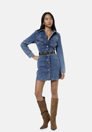 Vrouw die een denimjurk met knopen draagt met lange mouwen en een bruine riem, gecombineerd met bruine suède kniehoge laarzen, staand met hand in zij.