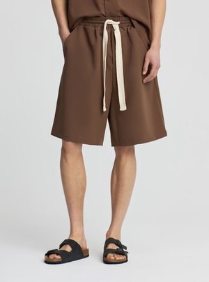 Uomo che indossa pantaloncini marroni al ginocchio con coulisse bianca e sandali neri aperti in punta, in piedi davanti a uno sfondo semplice.