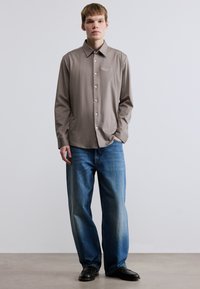 Μπεζ πουκάμισο με μακριά μανίκια και κουμπώματα, με διακριτική κεντημένη λεπτομέρεια στο στήθος, συνδυασμένο με μπλε τζιν φαρδιάς γραμμής και μαύρα παπούτσια.