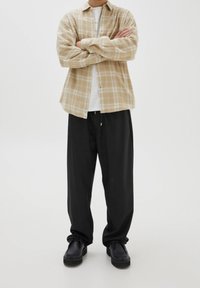 Hombre de pie con los brazos cruzados, vistiendo una camisa de cuadros beige, camiseta blanca, pantalones negros holgados y zapatos negros sobre un fondo blanco.