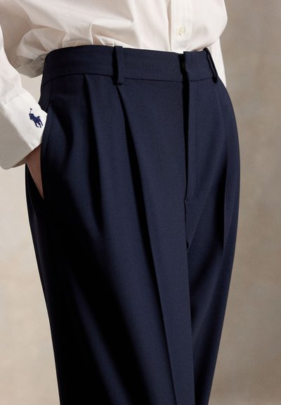 Navy tailored trousers with pleats, belt loops, and side pockets; fabric appears smooth with a slight sheen. White shirt cuffs visible.